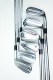 A set of six silver metal golf irons arranged diagonally on a clean white background, showing the club heads and portions of the shafts with black ferrule accents.